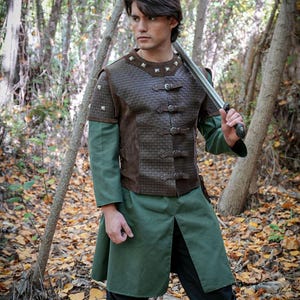Op de afbeelding: Een middeleeuwse outfit met een bruin leren vest met zilveren studs en gespen, gedragen over een groene tuniek en een shirt met lange mouwen. De outfit bevat een zwarte broek en bruine laarzen. Een zwaard wordt over de schouder gehouden. Het Rawblade-logo is zichtbaar.