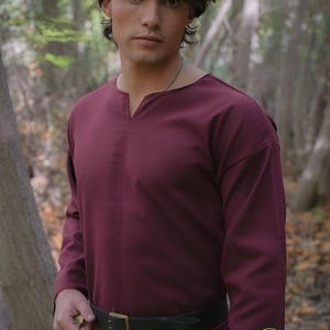 Peut inclure: Une tunique bordeaux à manches longues avec un col en V et une ceinture noire à boucle dorée. Le logo Rawblade est visible en bas à droite. La tunique est faite d'une matière douce et confortable.