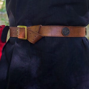 May include: A brown leather belt with a Celtic knot design and a metal buckle. The belt has a decorative metal piece with a Celtic knot design.