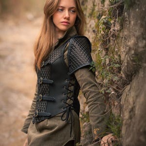 May include: A black leather vest with a quilted design and buckles. The vest has a laced-up front and shoulder pads. The vest is worn by a person standing in front of a stone wall.