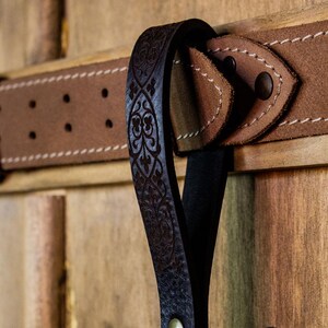 May include: A black leather belt with an intricate floral design. The belt has a brown leather buckle and is attached to a wooden surface.