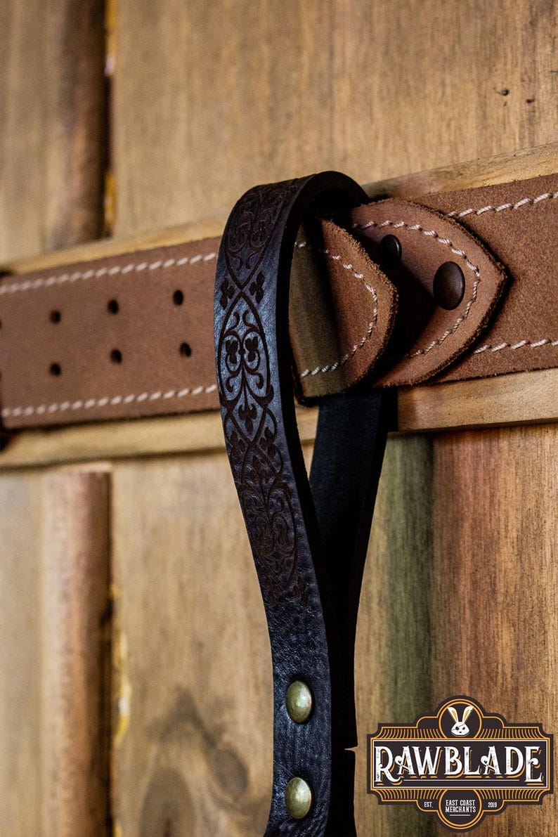 May include: A black leather belt with an intricate floral design. The belt has a brown leather buckle and is attached to a wooden surface.