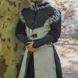 May include: A medieval-style tunic with a black and gray color scheme, featuring faux fur trim on the shoulders. The tunic has a leather-like texture and is fastened with ties and buckles. The Rawblade logo is visible in the bottom right corner.