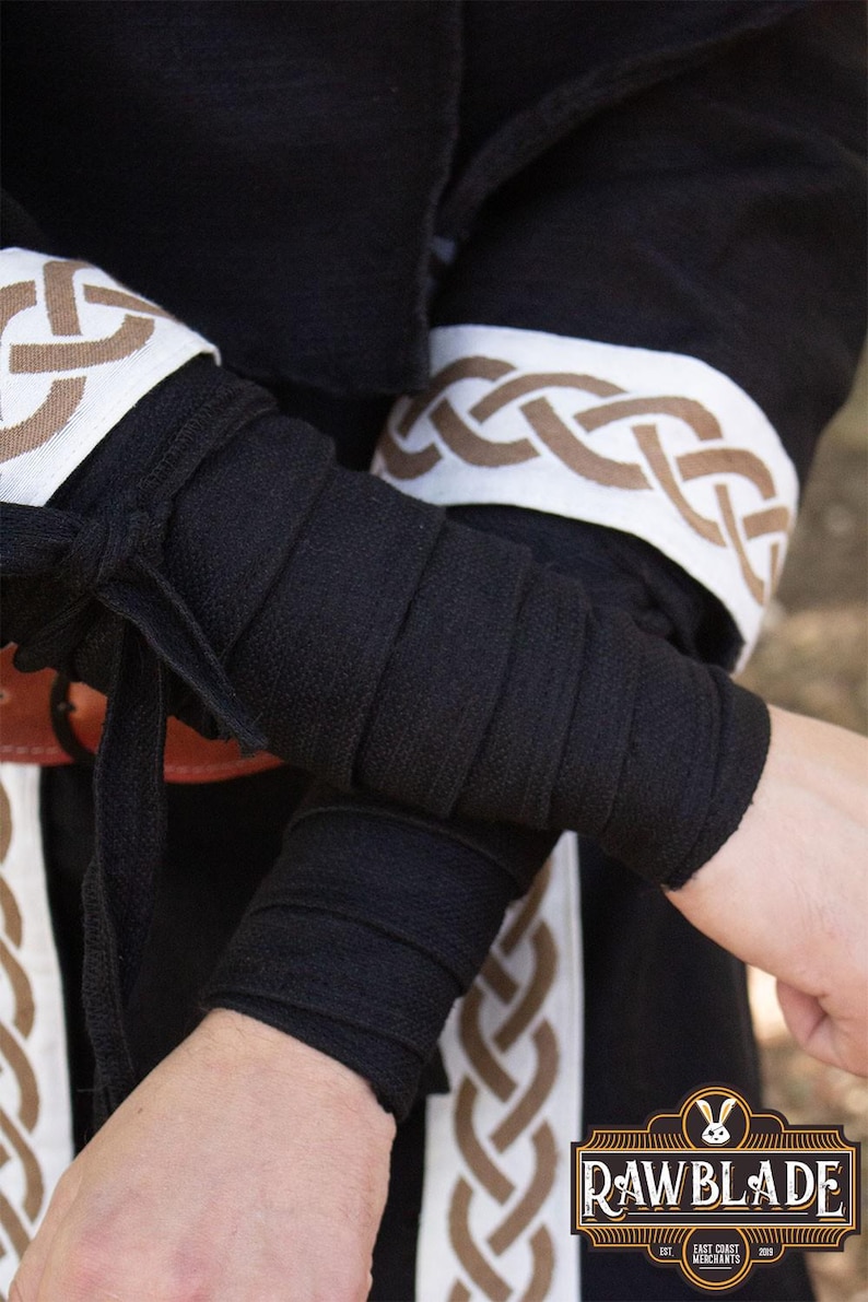 May include: Black fabric arm wraps with a white and brown braided design. The fabric is folded and tied at the wrist. The design features a repeating Celtic knot pattern.