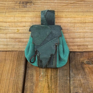 Puede incluir: Una pequeña bolsa de cuero verde azulado con una solapa de cuero verde oscuro y un cierre de cordón. La bolsa está decorada con costuras alrededor de los bordes.