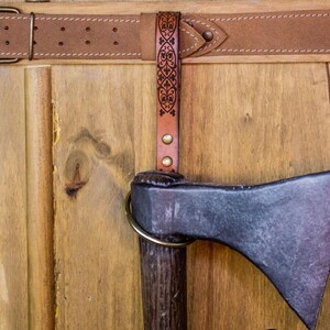 May include: A brown leather axe sheath with a decorative design hangs from a brown leather belt with a brass buckle. The sheath is attached to the belt with a brass rivet. The axe is not visible in the image.