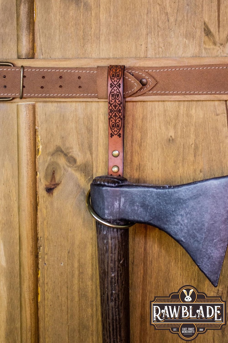 May include: A brown leather axe sheath with a decorative design hangs from a brown leather belt with a brass buckle. The sheath is attached to the belt with a brass rivet. The axe is not visible in the image.