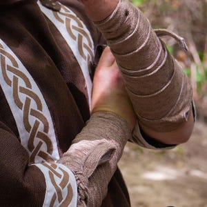 May include: A close-up of a person's arm wearing a brown leather arm guard with multiple straps. The arm guard is wrapped around the forearm and secured with a leather strap. The person is wearing a brown shirt with a white and brown Celtic knot pattern.