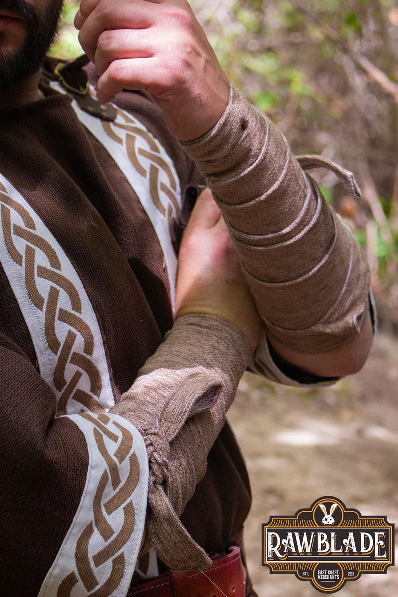 May include: A close-up of a person's arm wearing a brown leather arm guard with multiple straps. The arm guard is wrapped around the forearm and secured with a leather strap. The person is wearing a brown shirt with a white and brown Celtic knot pattern.