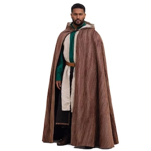 May include: A brown hooded cloak with a textured design. The cloak is worn over a green shirt, a cream-coloured scarf, and a brown belt. The outfit includes black trousers and boots. The cloak has a button closure at the neck.