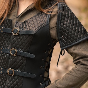 May include: Black leather armour vest with a quilted design and brass buckles. The vest has a lace-up back and is worn over a brown shirt. The vest has shoulder pads with a quilted design.