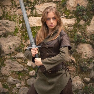 May include: A woman in a green tunic and brown leather armor holds a silver sword in front of a stone wall. The sword has a black handle and a silver blade. The woman is looking directly at the camera.