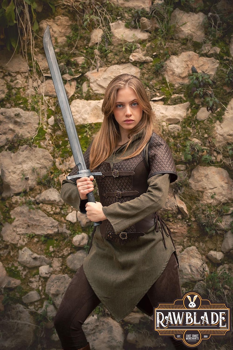 May include: A woman in a green tunic and brown leather armor holds a silver sword in front of a stone wall. The sword has a black handle and a silver blade. The woman is looking directly at the camera.
