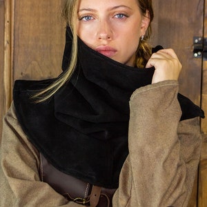 May include: A black hooded cowl made of soft fabric. The cowl is worn by a person with long blonde hair. The person is wearing a brown jacket over a brown leather vest. The background is a wooden wall.