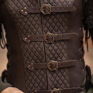 May include: A brown leather vest with a quilted diamond pattern and four adjustable straps with gold buckles. The vest has a laced back and is made by Rawblade East Coast Merchants.