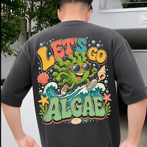 Könnte beinhalten: Ein anthrazitfarbenes T-Shirt mit einem Rückendruck, der eine grüne Algenfigur mit roter Sonnenbrille und Turnschuhen zeigt, die auf einer blauen Welle surft. Das Motiv enthält Muscheln, einen Seestern und den Schriftzug "LET'S GO ALGAE".