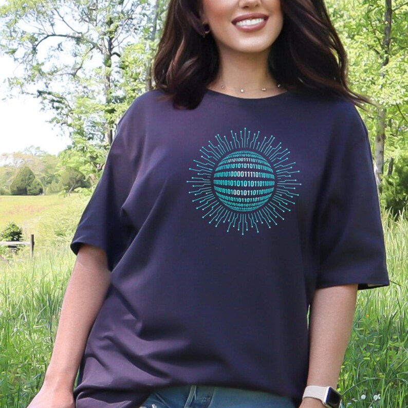Peut inclure: T-shirt bleu marine avec un motif de code binaire turquoise et blanc. Le motif est un cercle de code binaire avec des lignes rayonnantes. Le t-shirt a des manches courtes et une coupe d&eacute;contract&eacute;e. Le fond est un cadre naturel en ext&eacute;rieur.
