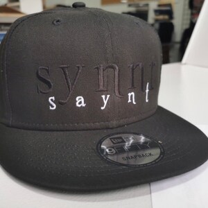 May include: A black and white baseball cap with the text "synt saynt" embroidered on the front. The cap has a black adjustable strap with a New Era logo and the text "9FIFTY SNAPBACK".