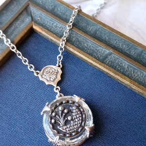 May include: A silver necklace with a clear, round pendant featuring a detailed design. The pendant hangs from a delicate chain. The necklace is displayed on a blue surface.