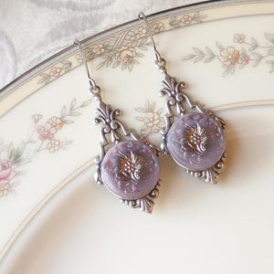 May include: A pair of vintage-style earrings. Each earring features a lavender-colored button with a gold-colored floral design, set in an ornate silver-toned frame. The earrings hang from delicate silver hooks, showcasing a classic, elegant design.