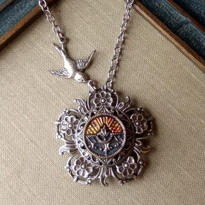 May include: Silver-toned necklace featuring a detailed pendant with a central design in shades of red, gold, and blue. The pendant is surrounded by floral embellishments. A small bird charm is attached to the chain. The necklace is displayed on a neutral background.