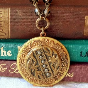 May include: Antique gold-tone locket necklace with a beaded chain. The round locket features floral and geometric designs, with a central oval adorned with small, round embellishments. The necklace rests on a stack of vintage books.
