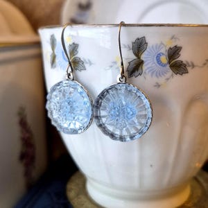 May include: A pair of round, light blue glass earrings featuring a pressed floral pattern and a scalloped edge. The circular glass pieces are suspended from silver-toned metal ear wires, creating a delicate and reflective accessory design.