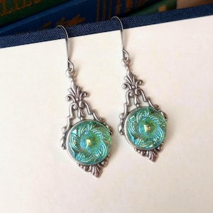 May include: A pair of silver-toned drop earrings. Each earring features a round, iridescent teal glass center with a swirling pattern, set within an ornate, filigree-style frame. The earrings have a hook closure.
