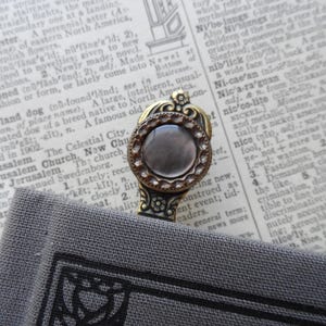 May include: A gold bookmark with a decorative design and a clear, round, iridescent stone in the center. The bookmark is resting on a book with a gray cover.