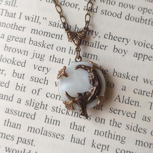 May include: A vintage-style necklace with a bronze-colored chain and pendant. The pendant features a white, circular mother-of-pearl base with a bronze crescent moon and stars design. The necklace is set against a backdrop of old book pages.