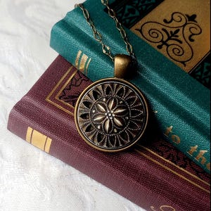 May include: A vintage-style pendant necklace with a detailed, circular design in antique brass tones. The pendant features a floral pattern with a central starburst design. The necklace rests on two antique books with gold accents and teal and burgundy covers.