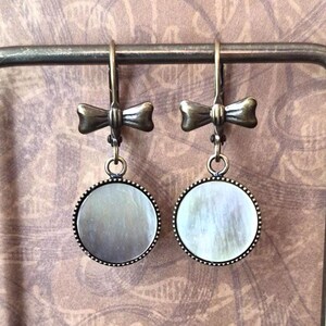 May include: A pair of drop earrings featuring a round, iridescent mother-of-pearl inlay set in a dark bronze-toned metal frame. Each earring is suspended from a lever-back closure adorned with a small, sculpted metal bow detail in a matching bronze finish.