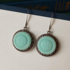 May include: A pair of vintage-style earrings. Each earring features a round, light blue center with a textured pattern, surrounded by a decorative, dark metal filigree frame. The earrings hang from simple, silver-toned hooks.