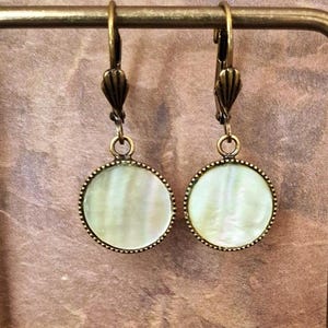 May include: A pair of drop earrings featuring round, iridescent white mother-of-pearl discs set in antique bronze-toned metal frames. The earrings have lever-back closures with a ribbed, fan-like design at the top of each hook.