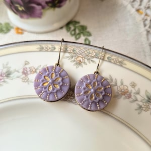 May include: A pair of round drop earrings featuring a lavender purple glass cabochon with a raised gold-toned floral pattern in the center and radiating lines. The earrings are suspended from simple bronze-toned metal hooks.