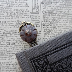 May include: A vintage-style bookmark with a decorative metal design and a purple and white button with a rhinestone in the center. The bookmark is lying on a gray book with black text.