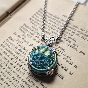 May include: A silver-toned necklace featuring a round, iridescent teal pendant with a raised, scalloped shell-like pattern. The pendant is held by a decorative silver metal setting with ornate filigree details and hangs from a twisted silver chain.