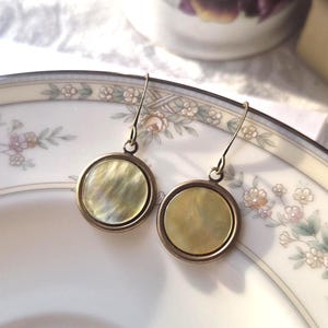 May include: A pair of drop earrings featuring circular, iridescent, pale yellow shell inlays set within antique bronze-toned metal frames. The earrings hang from simple curved metal hooks, showcasing a smooth, polished finish and a subtle, swirling texture on the surface of the round shell components.