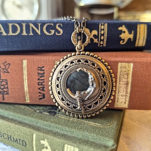 May include: A round bronze-toned pendant necklace featuring an intricate filigree border and a central relief of a figure seated on a crescent moon against a dark background. The pendant hangs from a matching bronze-toned metal chain link necklace.