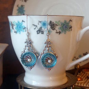 May include: A pair of silver-toned drop earrings with a round, light blue glass center, featuring a pearl and a swirl design. The earrings are displayed in front of a floral teacup.