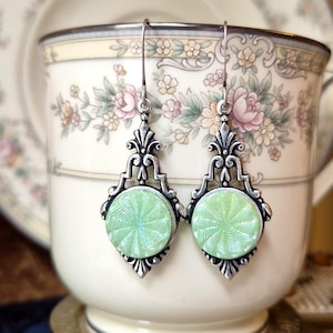 May include: A pair of drop earrings featuring round, light green glass cabochons with a swirling, sunburst-like texture. The cabochons are set in ornate, silver-toned metal frames with intricate scrollwork and hook-style ear wires for hanging.