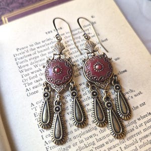 May include: A pair of dangle earrings featuring an antiqued brass finish with a central circular medallion in deep red enamel. Each earring has three hanging teardrop-shaped charms suspended from the base of the ornate metal medallion.