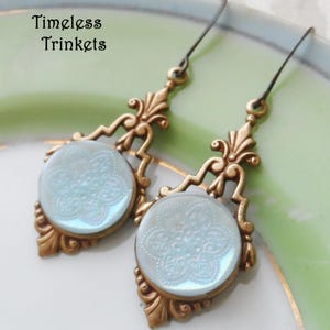 May include: A pair of dangle earrings with a light blue glass cabochon featuring a white floral design. The earrings have a gold-tone metal setting with a filigree design.