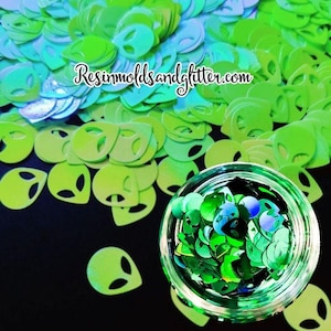 May include: A collection of green alien-shaped glitter confetti. The confetti is scattered across a black surface, with some contained in a clear glass jar. The website "Resinmoldsandglitter.com" is written in white cursive.