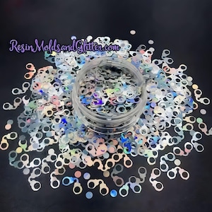 May include: A clear jar filled with iridescent silver glitter shaped like handcuffs. The glitter is scattered around the jar on a black background. ResinMoldsandGlitter.com is printed on the top of the image.
