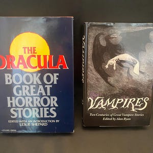 May include: Two vintage books: "The Dracula Book of Great Horror Stories" with a yellow sun graphic and red text, and "Vampires" with a dark illustration of a bat-like creature. Both are hardcover books.