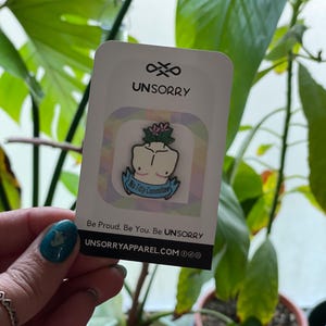 May include: A white card with the text "UNSORRY" in black. The card features a colourful illustration of a person with a flower on their head and the text "No Titty Committee". The card also includes the text "Be Proud. Be You. Be UNSORRY" and the website "UNSORRYAPPAREL.COM".