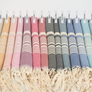 May include: A collection of Turkish towels in various colors, including dark gray, light gray, beige, mustard, coral, pink, damson, fuchsia, red, black, navy blue, petrol blue, sax blue, blue, turquoise, baby blue, and khaki green. The towels are rolled up and have fringe on the ends.