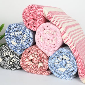 May include: A stack of rolled-up beach towels in various colors, including red, blue, pink, and black. The towels have a herringbone pattern and some have striped designs. The towels are made of a soft, absorbent material.