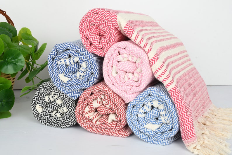 May include: A stack of rolled-up beach towels in various colors, including red, blue, pink, and black. The towels have a herringbone pattern and some have striped designs. The towels are made of a soft, absorbent material.